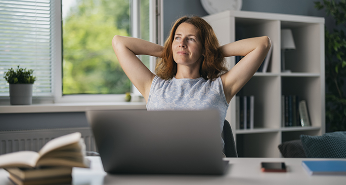 Woman at desk, in front of computer, with arms behind her head, looking out the window, thinking about her aspirations